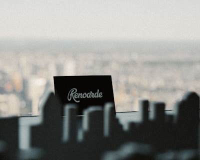 A black rectangular sign with the word 'Renovarde' written on it in white cursive font is placed against a blurred cityscape in the background. The foreground features the silhouette of what appears to be miniature buildings or parts of a city model.