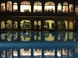 A well-lit building with arched windows overlooking a swimming pool at night. The upper floor appears to be a dining area with people seated at tables, while the lower floor seems to be a gym or fitness area with equipment visible. The pool in the foreground reflects the lights and arches from the building, creating a serene and somewhat luxurious atmosphere.