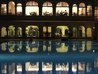 A well-lit building with arched windows overlooking a swimming pool at night. The upper floor appears to be a dining area with people seated at tables, while the lower floor seems to be a gym or fitness area with equipment visible. The pool in the foreground reflects the lights and arches from the building, creating a serene and somewhat luxurious atmosphere.