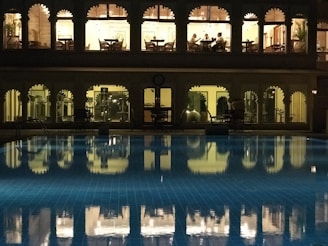 A well-lit building with arched windows overlooking a swimming pool at night. The upper floor appears to be a dining area with people seated at tables, while the lower floor seems to be a gym or fitness area with equipment visible. The pool in the foreground reflects the lights and arches from the building, creating a serene and somewhat luxurious atmosphere.