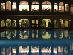A well-lit building with arched windows overlooking a swimming pool at night. The upper floor appears to be a dining area with people seated at tables, while the lower floor seems to be a gym or fitness area with equipment visible. The pool in the foreground reflects the lights and arches from the building, creating a serene and somewhat luxurious atmosphere.