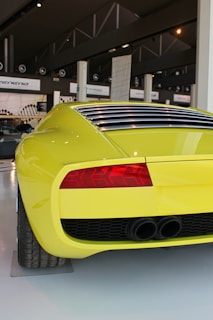 A bright yellow sports car is displayed in a modern showroom. The focus is on the rear end, showcasing the car's sleek design, detailed taillights, and dual exhaust system. The environment around the car is spacious, with signs of other vehicles and industrial design elements visible in the background.