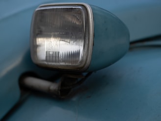 An old-fashioned car side mirror with an integrated rectangular headlight is attached to a blue vehicle. The surface shows signs of wear, with slight scratches and a vintage appearance.