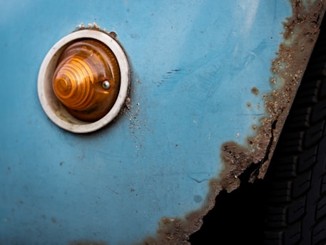 A close-up of a rusty, blue car body with an amber indicator light and visible corrosion along the edges.