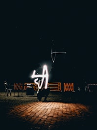 A nighttime scene with a dark background and a cobblestone floor reflecting light. In the center, a glowing light trails in the shape of the letter 'A'. Chairs and metal railings are faintly visible in the background.