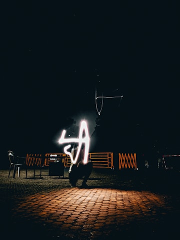 A nighttime scene with a dark background and a cobblestone floor reflecting light. In the center, a glowing light trails in the shape of the letter 'A'. Chairs and metal railings are faintly visible in the background.