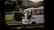 A white bus is captured in motion, driving along a road with a blurred background of greenery and hills. The bus appears to be empty except for the driver, and the light suggests it is either early morning or late afternoon.