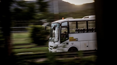 A white bus is captured in motion, driving along a road with a blurred background of greenery and hills. The bus appears to be empty except for the driver, and the light suggests it is either early morning or late afternoon.