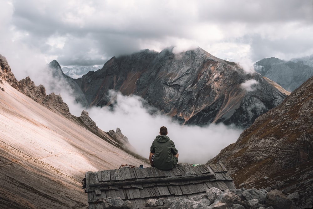 man in green jacket sitting on brown wooden fence looking at brown rocky mountain during daytime