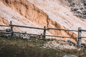 A rustic wooden fence stretches across a grassy area with scattered rocks, set against a backdrop of a steep, rocky cliff. The terrain appears dry and rugged, with a warm beige hue dominating the cliffside.