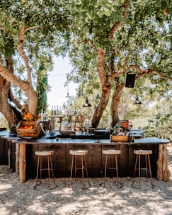 An outdoor bar setup is under the shade of large trees. The bar has a rustic wooden design with barstools made of wood and metal. Various bottles of alcohol and fresh fruit, including oranges in a wooden crate, are neatly arranged on the bar. Hanging lights and a speaker are suspended from the tree branches above. The surrounding area features gravel and is framed by greenery.