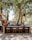 An outdoor bar setup is under the shade of large trees. The bar has a rustic wooden design with barstools made of wood and metal. Various bottles of alcohol and fresh fruit, including oranges in a wooden crate, are neatly arranged on the bar. Hanging lights and a speaker are suspended from the tree branches above. The surrounding area features gravel and is framed by greenery.