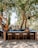 An outdoor bar setup is under the shade of large trees. The bar has a rustic wooden design with barstools made of wood and metal. Various bottles of alcohol and fresh fruit, including oranges in a wooden crate, are neatly arranged on the bar. Hanging lights and a speaker are suspended from the tree branches above. The surrounding area features gravel and is framed by greenery.
