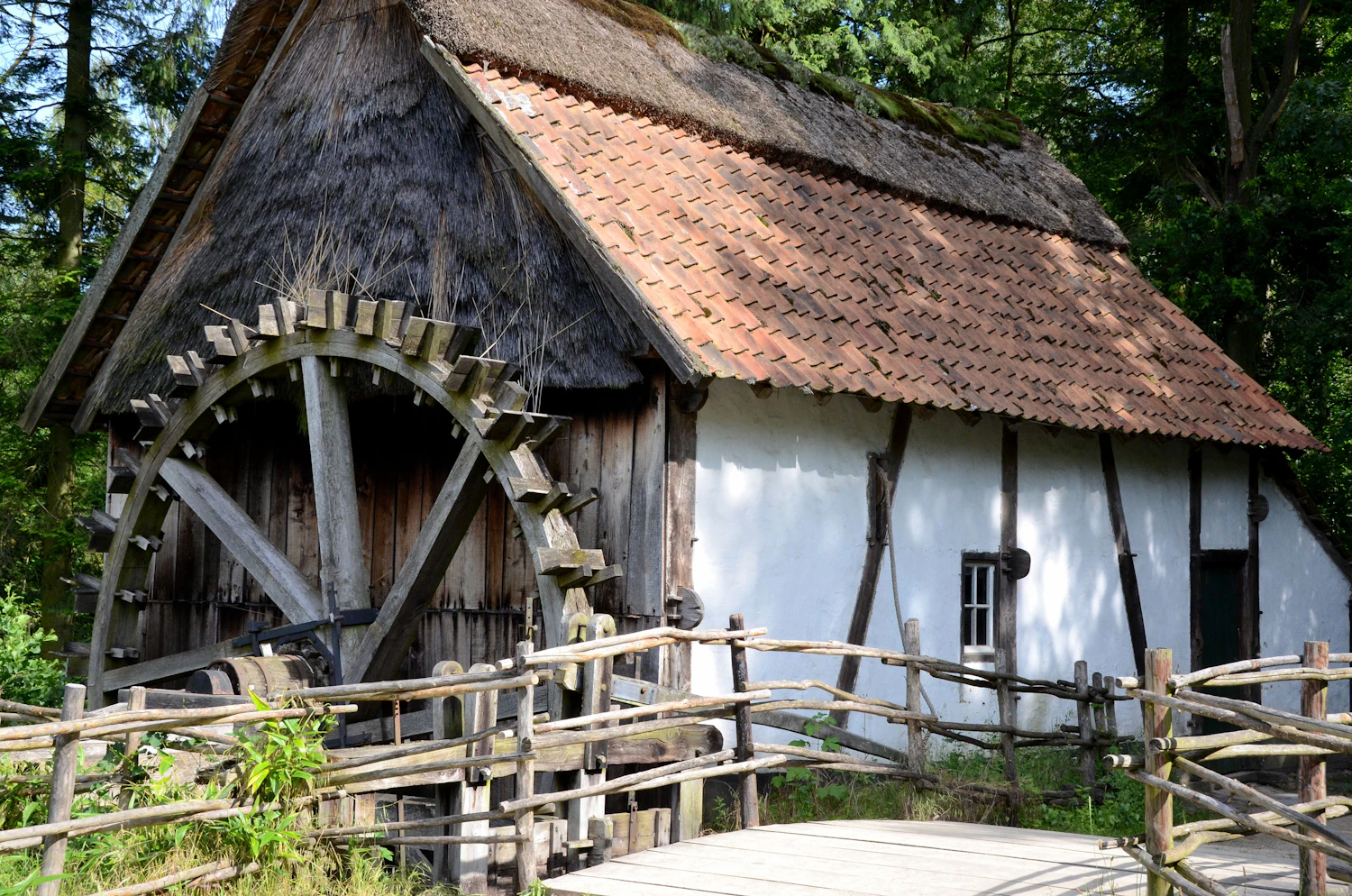 A white timber mill building with a large wooden wheel on its gable, seen from the yard