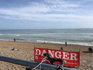 A beach scene with a clear blue sky and a calm ocean. People are seen on the shore and in the water, including a person in a kayak and swimmers. A red sign with the word 'DANGER' and warning messages is prominently displayed in front of the camera. A bicycle handlebar is partially visible at the bottom of the image.