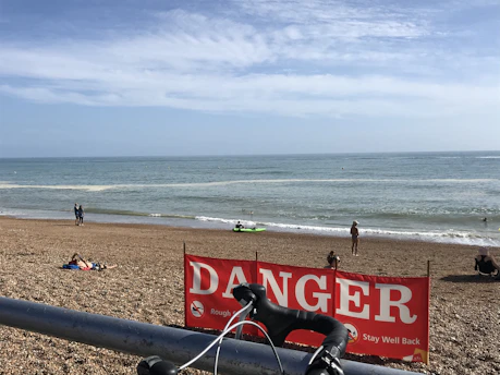 A beach scene with a clear blue sky and a calm ocean. People are seen on the shore and in the water, including a person in a kayak and swimmers. A red sign with the word 'DANGER' and warning messages is prominently displayed in front of the camera. A bicycle handlebar is partially visible at the bottom of the image.
