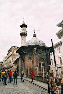 A bustling street scene with a historic mosque featuring intricate architecture and a towering minaret. People in colorful clothing, including a woman in a bright red coat, walk along the cobbled street. The sky is overcast, adding a subdued tone to the scene. A bird is captured mid-flight near the minaret.