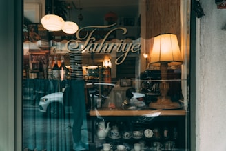 A cozy cafe interior is visible through a window, featuring a large, warmly lit lamp and shelves with various glassware and a clock. Bottles of syrup are displayed on a shelf, and vintage-style decorations add charm to the ambiance. The word 'Fahriye' is elegantly written on the window, reflecting a street scene including a white car.