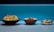 Three dishes are arranged side by side on a tiled surface with a vibrant blue background. The first dish on the left is a fresh salad featuring sliced radishes, greens, and various vegetables. The middle bowl contains a creamy soup or stew garnished with what appears to be herbs or small greens. The dish on the right is a dessert with layers of cream, cookie crumbs, and garnished with mint leaves.