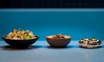 Three dishes are arranged side by side on a tiled surface with a vibrant blue background. The first dish on the left is a fresh salad featuring sliced radishes, greens, and various vegetables. The middle bowl contains a creamy soup or stew garnished with what appears to be herbs or small greens. The dish on the right is a dessert with layers of cream, cookie crumbs, and garnished with mint leaves.