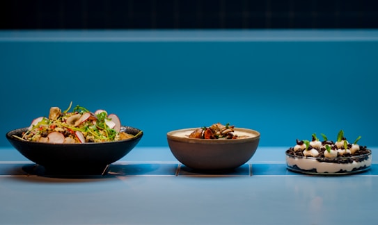 Three dishes are arranged side by side on a tiled surface with a vibrant blue background. The first dish on the left is a fresh salad featuring sliced radishes, greens, and various vegetables. The middle bowl contains a creamy soup or stew garnished with what appears to be herbs or small greens. The dish on the right is a dessert with layers of cream, cookie crumbs, and garnished with mint leaves.