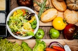 A vibrant display of fresh ingredients including a bowl of mixed salad with leafy greens, tomatoes, and bell peppers. Surrounding the bowl are various types of bread and fresh vegetables such as tomatoes, carrots, and onions. Sprigs of rosemary add a touch of greenery, while the rustic wooden surface enhances the natural theme.