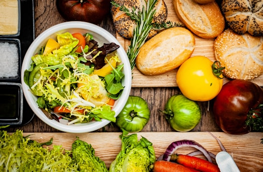 A vibrant display of fresh ingredients including a bowl of mixed salad with leafy greens, tomatoes, and bell peppers. Surrounding the bowl are various types of bread and fresh vegetables such as tomatoes, carrots, and onions. Sprigs of rosemary add a touch of greenery, while the rustic wooden surface enhances the natural theme.