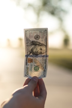 A hand holds a crumpled US hundred-dollar bill against a blurred outdoor background with soft sunlight. The focus is on the bill, while the background appears serene and out of focus.