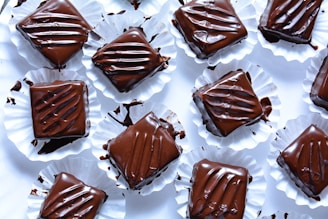 Several pieces of chocolate dessert are arranged on a white surface. Each piece is square-shaped, coated in glossy dark chocolate, and placed in crinkled white paper cups. The chocolate topping appears smooth with artistic drizzle patterns, and some smudges of chocolate can be seen on the cups.