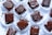 Several pieces of chocolate dessert are arranged on a white surface. Each piece is square-shaped, coated in glossy dark chocolate, and placed in crinkled white paper cups. The chocolate topping appears smooth with artistic drizzle patterns, and some smudges of chocolate can be seen on the cups.