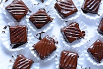 Several pieces of chocolate dessert are arranged on a white surface. Each piece is square-shaped, coated in glossy dark chocolate, and placed in crinkled white paper cups. The chocolate topping appears smooth with artistic drizzle patterns, and some smudges of chocolate can be seen on the cups.