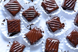 Several pieces of chocolate dessert are arranged on a white surface. Each piece is square-shaped, coated in glossy dark chocolate, and placed in crinkled white paper cups. The chocolate topping appears smooth with artistic drizzle patterns, and some smudges of chocolate can be seen on the cups.