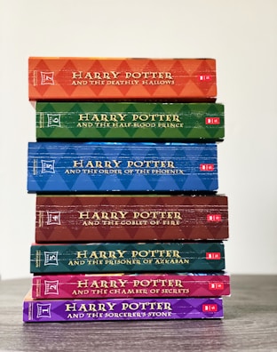 A neatly stacked collection of seven Harry Potter books, each with a uniquely colored spine. The spines display the titles, arranged in reverse chronological order, starting from 'Harry Potter and the Deathly Hallows' on top to 'Harry Potter and the Sorcerer's Stone' at the bottom. The spines exhibit a textured, patterned design with bold lettering.