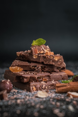 A stack of chocolate pieces, adorned with almonds and a fresh mint leaf on top, scattered with chocolate shavings and nuts around on a dark background.