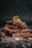 A stack of chocolate pieces, adorned with almonds and a fresh mint leaf on top, scattered with chocolate shavings and nuts around on a dark background.