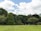 A lush, green landscape with a well-manicured lawn in the foreground. The scene is dominated by tall, dense trees with vibrant, leafy canopies under a partly cloudy sky.