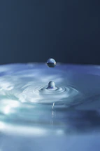 A close-up of a clear water droplet falling into a calm pool, reflecting soft natural light.