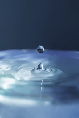 A close-up of a clear water droplet falling into a calm pool, reflecting soft natural light.