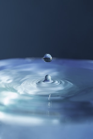 Close-up of a clear water droplet falling into a calm blue pool, reflecting purity.