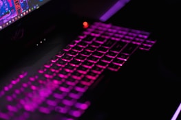An illuminated keyboard with backlit keys in shades of purple and pink against a dark background. The keys are visibly glowing, suggesting a gaming or colorful aesthetic. Part of a laptop screen is visible, showing a colorful interface.