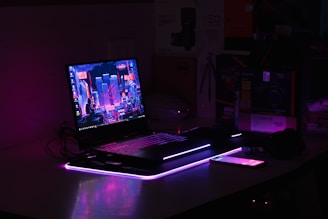 A dimly lit setup features a laptop with a vibrant, futuristic cityscape displayed on its screen. The scene is enhanced with ambient purple and pink LED lighting, casting a glowing effect on the surroundings. An illuminated smartphone lies nearby, adding to the tech-savvy atmosphere. Various boxes and technological gadgets are visible in the background.