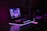 A dimly lit setup features a laptop with a vibrant, futuristic cityscape displayed on its screen. The scene is enhanced with ambient purple and pink LED lighting, casting a glowing effect on the surroundings. An illuminated smartphone lies nearby, adding to the tech-savvy atmosphere. Various boxes and technological gadgets are visible in the background.