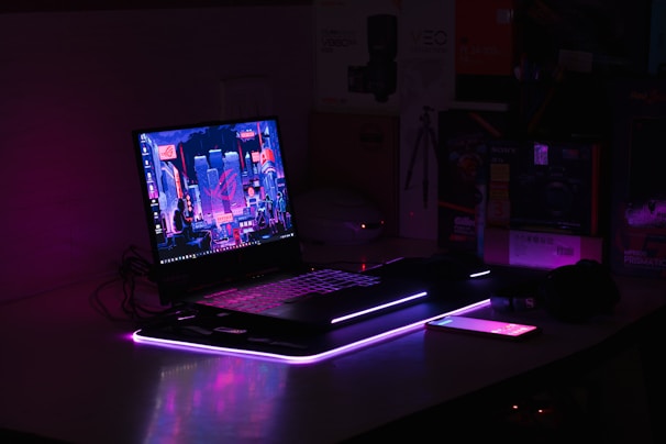 A dimly lit setup features a laptop with a vibrant, futuristic cityscape displayed on its screen. The scene is enhanced with ambient purple and pink LED lighting, casting a glowing effect on the surroundings. An illuminated smartphone lies nearby, adding to the tech-savvy atmosphere. Various boxes and technological gadgets are visible in the background.