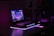 A dimly lit setup features a laptop with a vibrant, futuristic cityscape displayed on its screen. The scene is enhanced with ambient purple and pink LED lighting, casting a glowing effect on the surroundings. An illuminated smartphone lies nearby, adding to the tech-savvy atmosphere. Various boxes and technological gadgets are visible in the background.