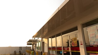 A convenience store exterior with large glass windows reflecting the warm afternoon light. The building features a flat roof and contains various signage and advertisements on the doors. Yellow bollards and a small parking area can be seen in front, with a motorcycle and rider wearing a helmet nearby.
