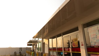 A convenience store exterior with large glass windows reflecting the warm afternoon light. The building features a flat roof and contains various signage and advertisements on the doors. Yellow bollards and a small parking area can be seen in front, with a motorcycle and rider wearing a helmet nearby.
