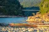 A serene riverside scene with several people engaging in leisurely activities on a rocky shore. Wooden boats are moored along the water's edge. The lush green forest and hills form a backdrop, with a steel bridge spanning the river in the distance.
