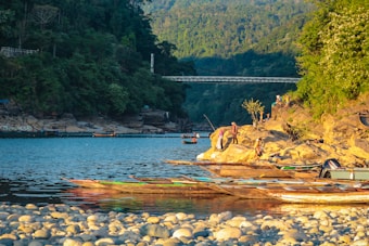 A serene riverside scene with several people engaging in leisurely activities on a rocky shore. Wooden boats are moored along the water's edge. The lush green forest and hills form a backdrop, with a steel bridge spanning the river in the distance.