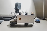 A detailed miniature model of a UPS delivery truck sits on a desk, with various office items in the background. The truck features the UPS logo and is accompanied by a smaller blue and white toy vehicle.