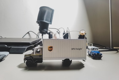 A detailed miniature model of a UPS delivery truck sits on a desk, with various office items in the background. The truck features the UPS logo and is accompanied by a smaller blue and white toy vehicle.
